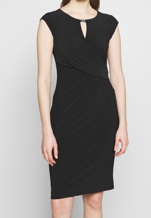 Robe noire sans manches jusqu'aux genoux avec un design portefeuille, encolure en trou de serrure avec une petite attache, portée par une femme debout.