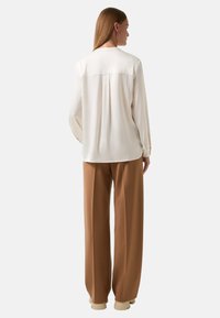 Crème blouse met lange mouwen en een plissé achteraan, gecombineerd met bruine wijdelijf broeken en beige schoenen. Eenvoudig ontwerp, gladde textuur.