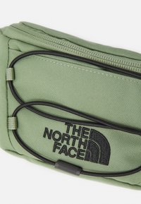 Sac banane en tissu vert avec fermeture éclair noire, broderie de logo et détails en cordon élastique. Présente une surface texturée et une forme courbée.