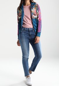 Blue velour bomber jacket with colorful floral patterns, paired with a pink t-shirt and medium-wash skinny jeans. White sneakers complete the look.