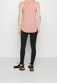 Light pink sleeveless top with a curved hem and asymmetrical back detail, paired with black leggings and black shoes featuring a gold accent.