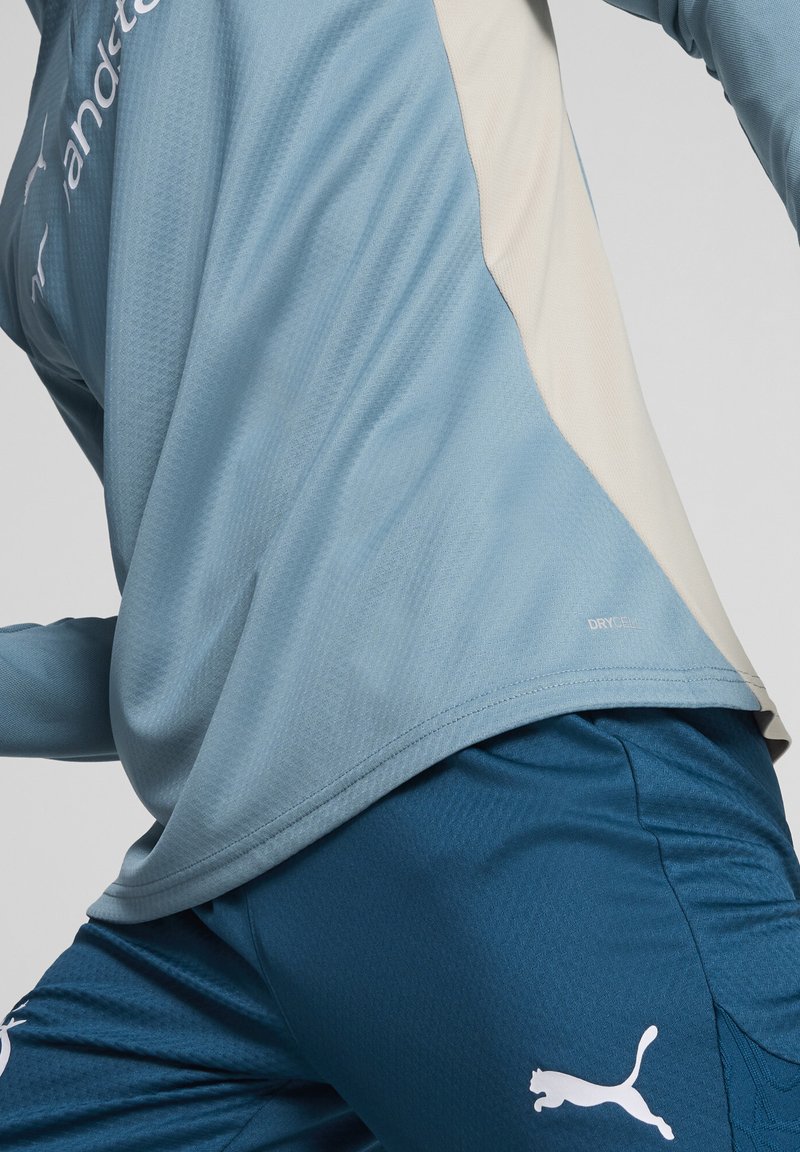 La maglietta sportiva blu presenta un tessuto testurizzato con dettagli crema; abbinata a pantaloni corti blu scuro che mostrano un logo bianco e dettagli di design sportivo.