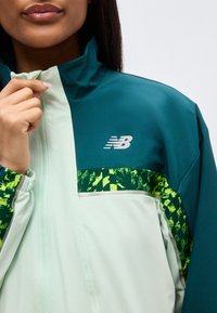 Γυναίκα που ρυθμίζει το φερμουάρ σε τζάκετ New Balance σε τυρκουάζ και ανοιχτό πράσινο με μοτίβο από νέον πράσινο στο μανίκι και το στήθος.