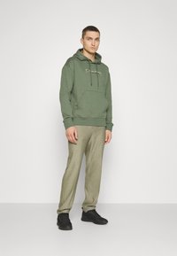 Felpa verde oliva con tasca a marsupio e cordino, abbinata a pantaloni leggeri a quadretti e sneakers nere. Design minimalista.