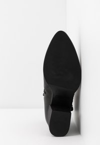 Botte noire à bout pointu avec semelle texturée et talon bloc, positionnée à la verticale contre un mur blanc d'angle.
