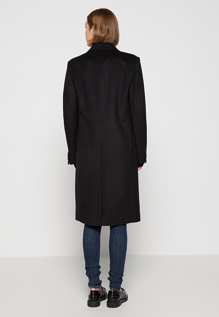 Manteau noir en laine avec une silhouette longue, col à revers et poches latérales. Associé à un jean sombre et des chaussures noires.