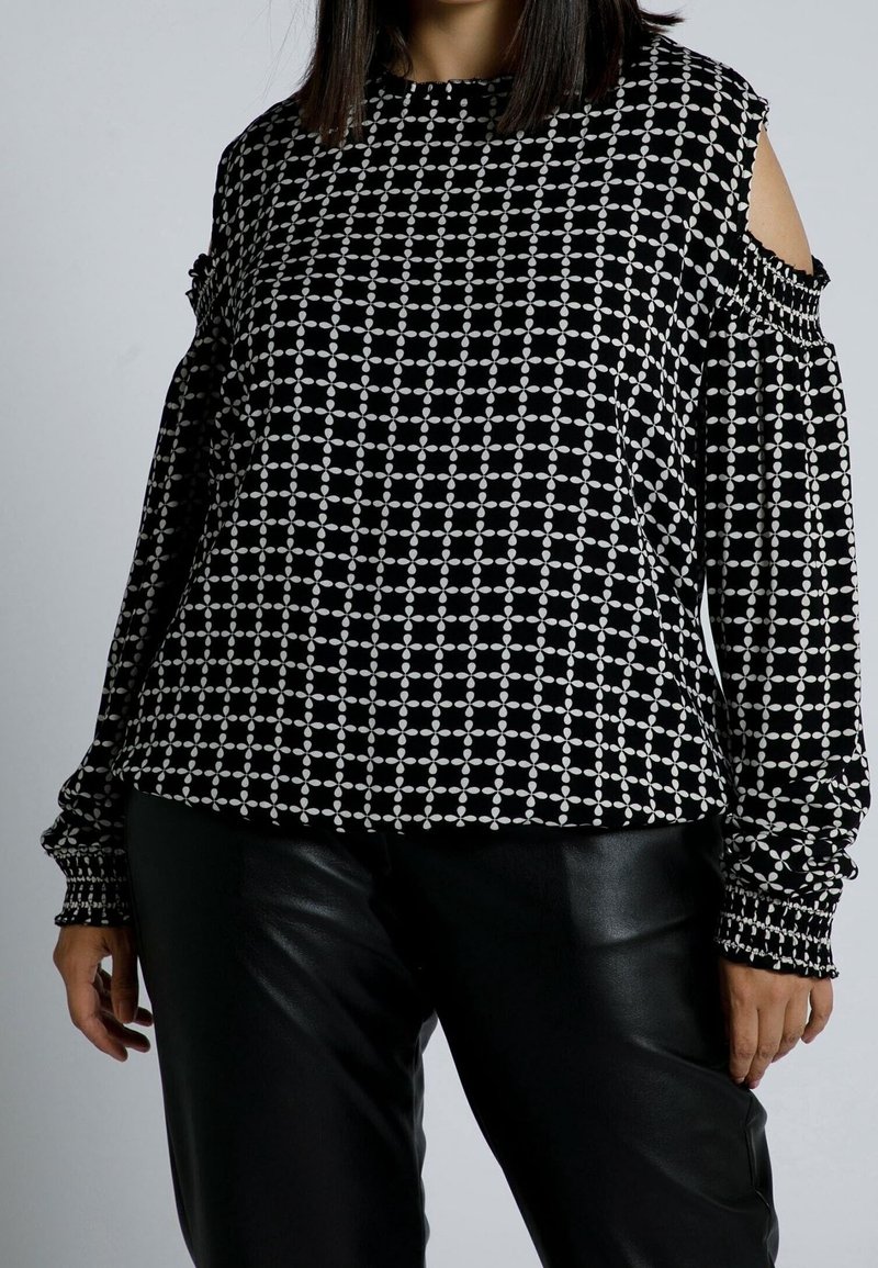 Schwarz-weiß gemustertes Oberteil mit Off-Shoulder-Details, langen Ärmeln und gerafften Manschetten, aus weichem Stoff gefertigt. Kombiniert mit schwarzen Hosen.