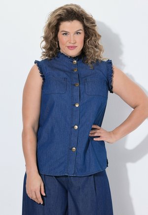 Kobieta o kręconych włosach, ubrana w niebieską bezrękawnikową koszulę na guziki z denimu oraz pasujące szerokie spodnie, stoi z jedną ręką na biodrze.
