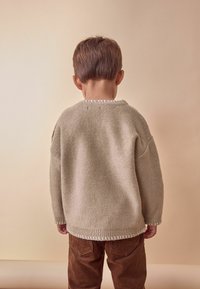 Beige neulé tröja med ribbad nederkant och texturerad yta, parat med bruna kordflodbyxor. Bakgrunden är en dämpad beige.