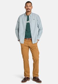 Světle modrá košile na knoflíky přes zelené tričko, doplněná béžovými cargo kalhotami. Hnědé šněrovací boty mají robustní design.