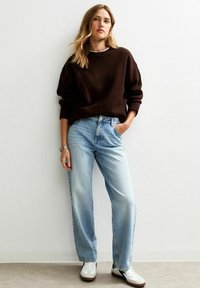 Suéter de punto marrón, jeans de corte relajado azul claro, zapatillas blancas con acentos negros, de pie contra un fondo neutro.