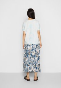 Ljusblå tie-dye t-shirt i kombination med en lång blommig kjol i blå, gräddvit och gyllene mönster; bärs med svarta slip-on sandaler.