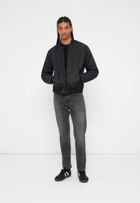 Sort quiltet bomberjakke med ribbede manchetter og kant, båret over en sort skjorte. Grå jeans og sorte sneakers med hvide striber fuldender outfittet.