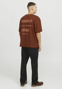 Camiseta oversized marrón con texto en la parte posterior, combinada con pantalones negros y zapatos marrones. La tela parece suave, con un ajuste relajado y mangas cortas.