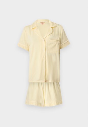Jeune femme debout pieds nus portant une chemise crème à manches courtes avec col et un short assorti sur un fond blanc uni.