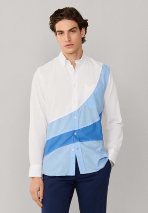 Hombre vistiendo una camisa blanca de manga larga con rayas diagonales azules, pantalones azul marino y zapatos marrones sin cordones, de pie frente a un fondo liso.