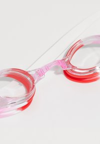 Lunettes de natation transparentes avec des garnitures en silicone rose et rouge et un pont nasal réglable rose sur une surface blanche.