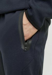 Les joggers de sport navy sont dotés d'une poche latérale avec une fermeture éclair noire et un tissu texturé, mettant en avant un design moderne et fonctionnel.