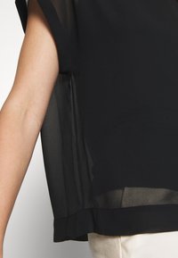Schwarze Bluse mit transparenter Textur, lockerer Passform und abgerundetem Saum. Die Ärmel zeigen eine leichte Transluzenz. Einfaches, unauffälliges Design.