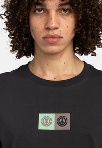 Schwarzes Baumwoll-T-Shirt mit zwei rechteckigen Designs: Ein hellgrünes Blattlogo auf der linken Seite und ein dunkelbraunes Friedenslogo auf der rechten Seite.