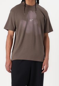 Hnědé bavlněné tričko s uvolněným střihem, na hrudi má nenápadné logo Nike a vybledlý vzor v světlejším odstínu.