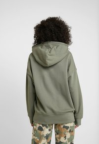 Olive green hoodie with a large, adjustable hood, soft texture, relaxed fit, and ribbed cuffs; worn with camo-patterned pants.