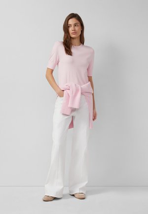 Frau steht in weißen, weit geschnittenen Hosen, einem hellrosa kurzärmeligen Oberteil und einem rosa Pullover, der um die Taille gebunden ist, und trägt beigefarbene Schuhe.