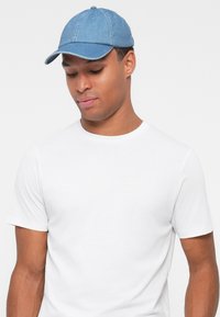 Blaues Denim-Baseballcap mit einem gebogenen Schirm, sechs Paneelen und dekorativen Nähten. Trägt ein schlichtes weißes T-Shirt.