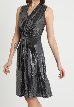 Mouwloze zwarte en zilveren paillettenjurk met een geplooide draaiding aan de taille, gedragen door een persoon met zichtbare armtatoeages.
