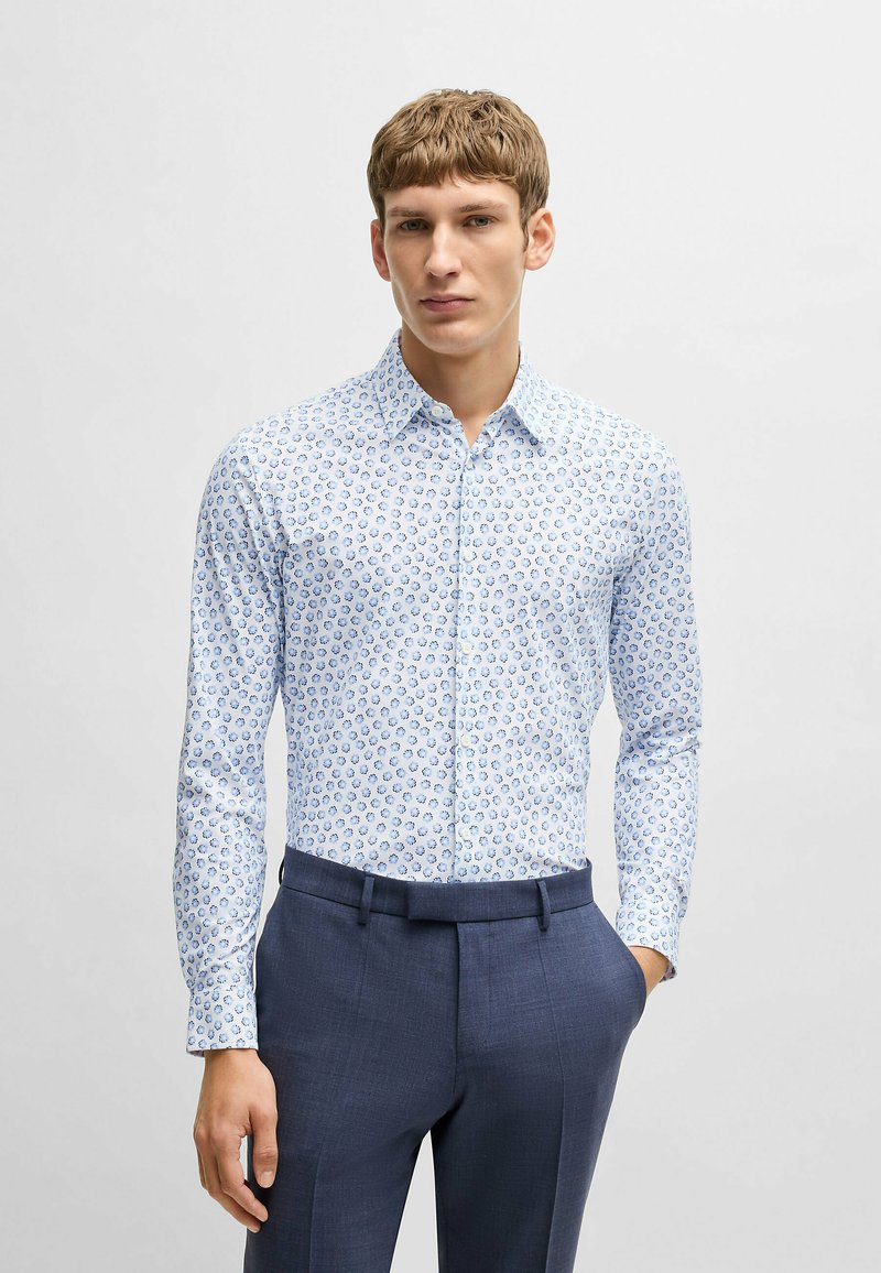 Chemise blanche à manches longues avec un motif floral bleu, associée à un pantalon tailleur bleu foncé. La chemise dispose d'un col boutonné.