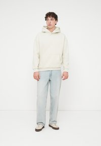 Κρεμ φούτερ με κουκούλα και τσέπη μπροστά, συνδυασμένο με ανοιχτόχρωμο τζιν και μπεζ αθλητικά παπούτσια. Απλός, άνετος σχεδιασμός σε χαλαρή γραμμή.