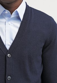 Mörkblå cardigan i stickad kvalité med V-ringning, fyra knappar och en mjuk ytstruktur, som är överlagd på en ljusblå skjorta.