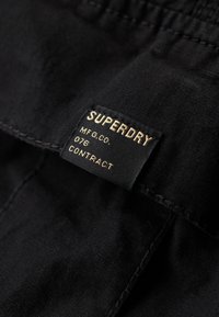 Etiqueta de tela negra con texto dorado que dice "SUPERDRY MFG.CO. 076 CONTRACT." Textura suave, forma rectangular, bordes cosidos.