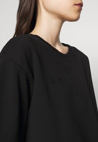 Woman with long dark hair wearing a black sweatshirt with subtle embroidered text "MARGIELA 6" on chest, viewed from shoulder up.