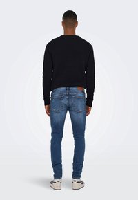 Sudadera negra, jeans ajustados azul y zapatillas blancas. Vista trasera cercana que resalta las costuras, la textura del denim y los detalles de la cinturilla.