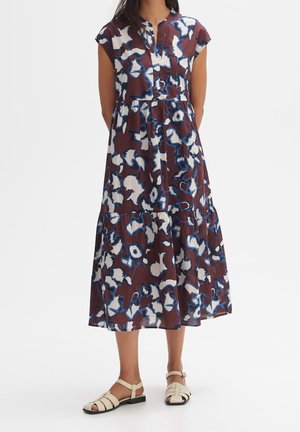 Frau trägt ein ärmelloses, knöpfenverschluss Midi-Kleid mit einem abstrakten Blumenmuster in Blau, Weiß und Braun sowie weiße flache Sandalen.