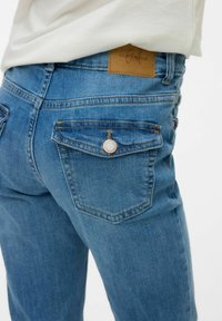 Pantalones vaqueros de mezclilla azul con una altura media, que presentan dos bolsillos traseros con solapas puntiagudas y una etiqueta de cuero marrón en la cinturilla.