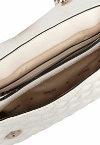 Doorstikte witte handtas met gouden hardware, ritssluitingvak, zachte binnenvoering en merknaam "GUESS" zichtbaar aan de binnenkant.