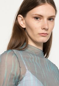 Top transparente de manga longa com riscas onduladas em azul e verde sobre um fundo cinza, com um decote alto e detalhes de costura delicados.