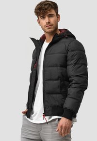 Schwarze Pufferjacke mit maroonem Innenfutter, ausgestattet mit einem Reißverschluss vorne, Seitentaschen, elastischen Bündchen und roten Kordeln an der Kapuze.