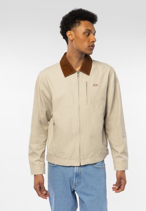Ung mann i beige jakke med brun krage og blå jeans, ser mot høyre mot en ensfarget lys bakgrunn.