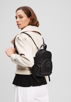 Schwarzer Nylon-Rucksack mit runder Form, vorderem Reißverschluss und eingeprägtem Logo. Auf dem Rücken getragen, kombiniert mit einem beigen Trenchcoat und einem schwarzen Rüschenshirt.