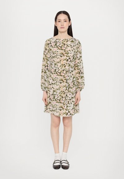 Robe florale en beige clair avec des motifs rose et vert, manches bouffantes longues, taille cintrée et ourlet au-dessus du genou. Portée avec des chaussures sombres et des chaussettes blanches.