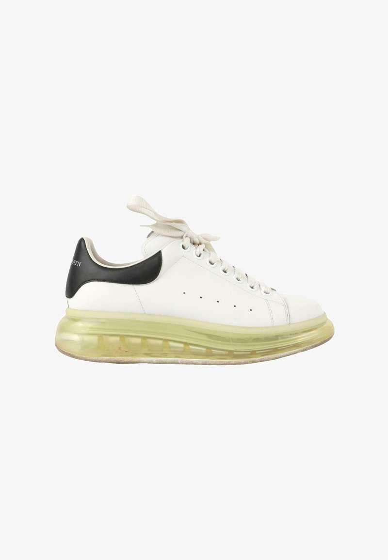 Baskets blanches avec languette noire au talon, semelle jaune translucide épaisse et lacets blancs noués, vues de l’extérieur.