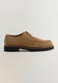 Chaussure en cuir suédé marron avec un bout arrondi, des coutures décoratives et une semelle en caoutchouc noir épaisse avec un motif de préhension texturé.