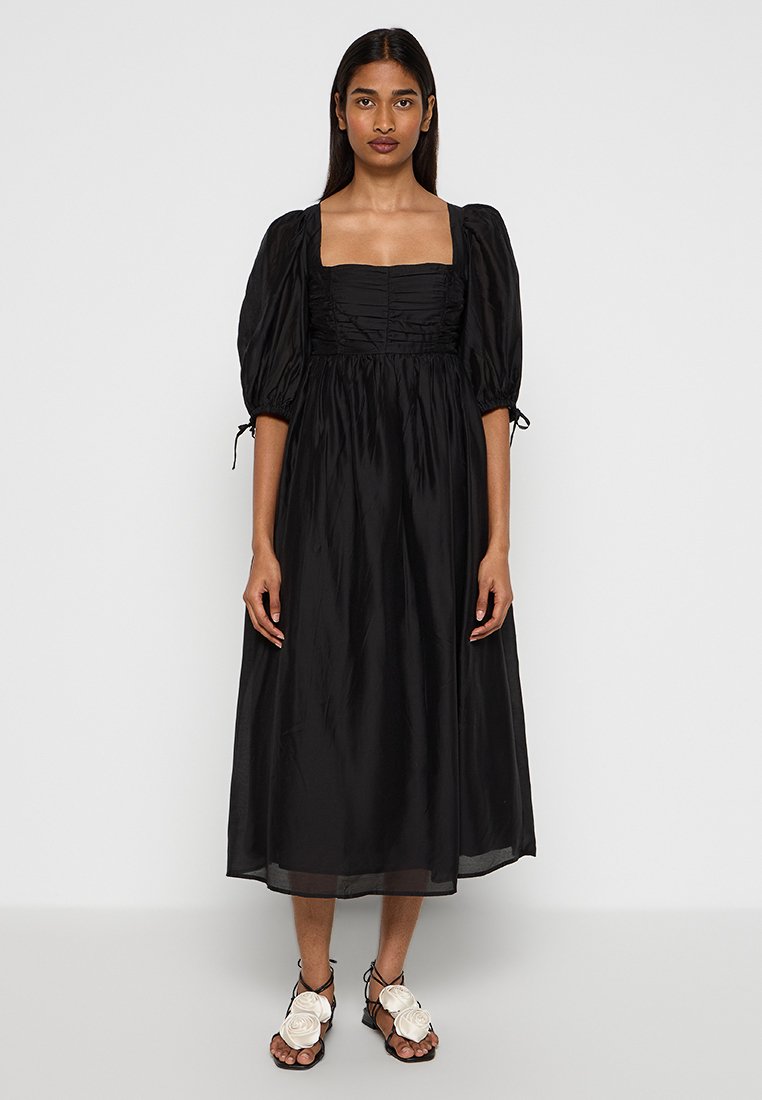 Robe midi noire avec manches bouffantes et décolleté carrée. Fabriquée en tissu lisse avec des détails froncés au niveau du corsage. Accents et liens aux manches.