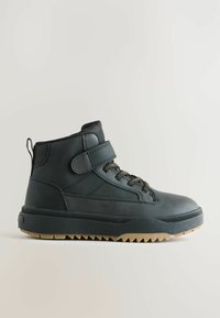 Svart mid-top stövlar med en slät ovansida, texturerade sidor, ett kardborreband och en kontrasterande tan-gummisul med ett sågad mönster.