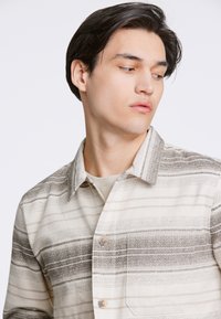 Camicia leggera a righe nei toni beige e grigi. Caratterizzata da colletto, bottoni frontali e un motivo testurizzato.