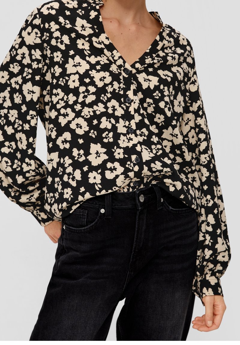 Femme portant un chemisier noir à boutons avec un imprimé floral beige et un jean noir taille haute, debout avec une main dans la poche.