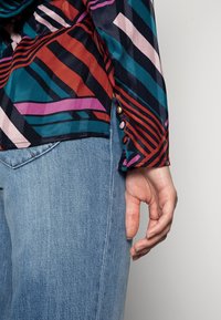Blouse colorée avec de larges rayures en sarcelle, orange, rose et noir. Présente un détail de bouton sur la manche, accompagnée de jeans bleu clair.